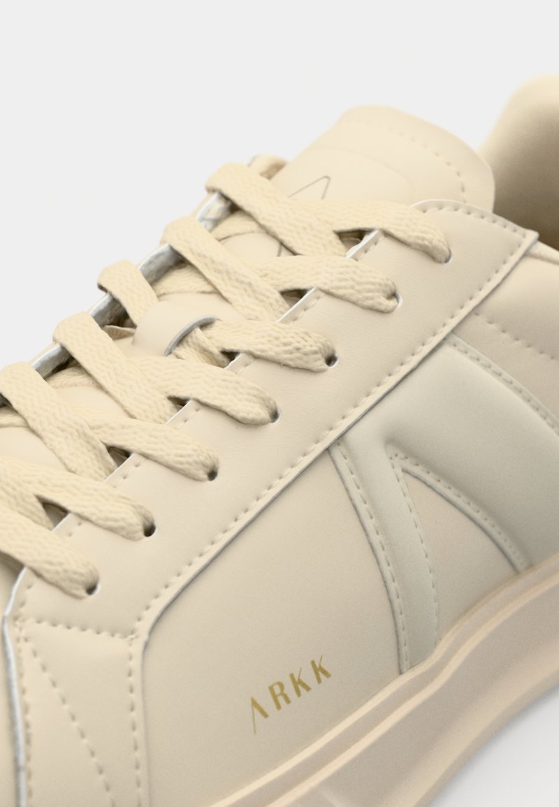 Zapatilla beige con un acabado de cuero suave, que presenta cordones planos, un cuello acolchado y un detalle de logo dorado en el costado.