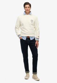 Sudadera de color crema claro con puños acanalados, utilizada sobre una camisa de botones azul. Jeans oscuros y zapatillas beiges completan el atuendo.