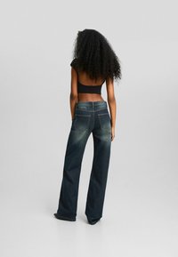 Kvinna med långt lockigt hår, iklädd en svart kort topp och löst sittande mörkblå jeans, står mot en enfärgad bakgrund med ryggen vänd mot kameran.