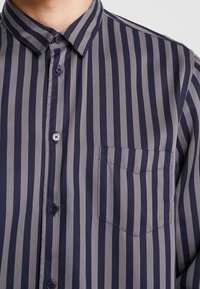 Camicia a righe navy e grigia con bottoni, dotata di taschino, colletto classico e texture liscia. Bottoni dello stesso colore del tessuto.
