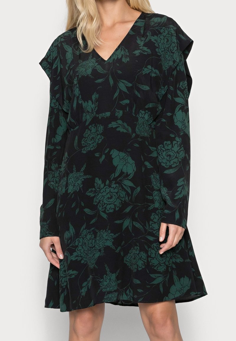 Robe noire à manches longues avec imprimé floral vert, col en V et détail subtil de volants aux épaules portée par une personne debout.