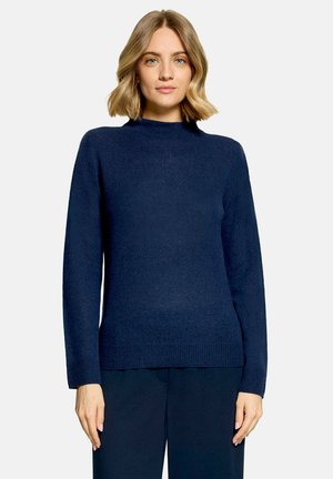 Femme aux cheveux blonds mi-longs, portant un pull bleu foncé à manches longues et un pantalon bleu foncé assorti, debout sur un fond uni.