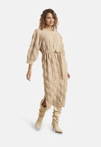 Donna che cammina indossando un vestito midi beige in maglia texturizzata con cintura e spacchi, abbinato a stivali alti color crema morbidi con tacco, su sfondo bianco.