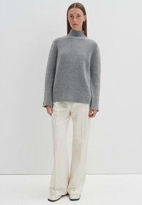 Grau gestrickter Pullover mit hohem Kragen und langen Ärmeln, kombiniert mit weißen weit geschnittenen Hosen und schwarzen quadratischen Schuhen.