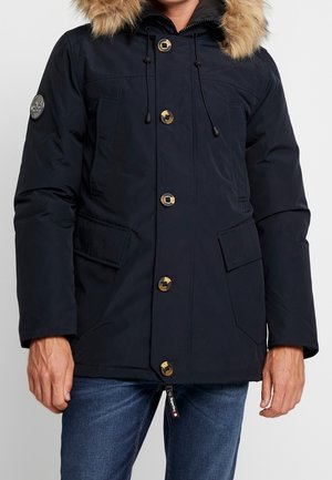 Homme portant un manteau d'hiver bleu marine foncé avec une capuche en fourrure, de gros boutons, des poches avant à rabat et un jean bleu.