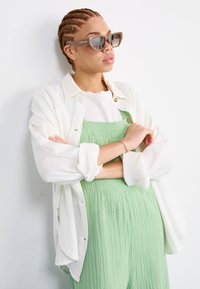 Witte, lichte, lange mouwen shirt over een groene, gestructureerde overall met knopen. Bruine zonnebril met een rechthoekige vorm. Neutrale achtergrond.
