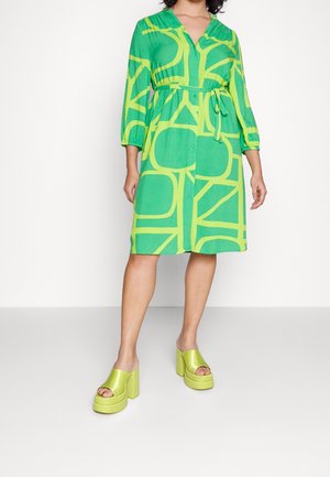 Vrouw draagt een groen jurk tot op de knie met gele geometrische patronen, lange mouwen en een taillekoord, gecombineerd met limegroene platform sandalen.