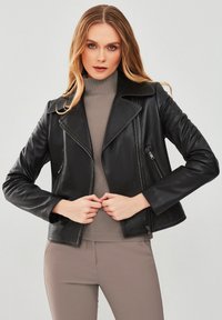 Zwarte leren motorjack met een gegroefde revers, asymmetrischerits en voorzakken. Gecombineerd met een grijze coltrui en strakke beige broek.