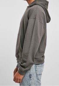 Felpa grigia in cotone con tasca frontale, cappuccio e polsini a costine. Indossata con jeans azzurri strappati, mostrata di lato.