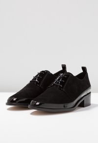 Scarpe oxford nere in suede con punta e tacco in vernice lucida. Presentano lacci e una linguetta sul retro per facilitarne l'indossamento.