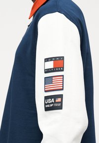 Μπλε και λευκό μανίκι μακρυμάνικου πουκάμισου με τρεις σημαίες: λογότυπο Tommy Hilfiger, αμερικανική σημαία και έμβλημα της ομάδας USA SailGP.