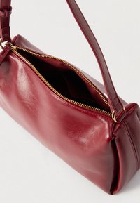 Sac d'épaule en cuir rouge avec une texture lisse, une fermeture éclair dorée et une bandoulière courte. Le sac a une forme rectangulaire et une ouverture supérieure.