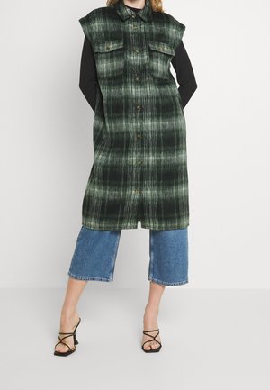 Kobieta ubrana w bezrękawnik zielony długi kamizel z kratą na czarnym topie, skrócone niebieskie dżinsy oraz czarne sandały na obcasie z paskami, stoi z rękami za plecami.