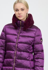Junge Frau in einer lila glänzenden Steppjacke mit einem dunkel-lila Pelzkragen und einem goldenen Reißverschluss, die frontal vor einem schlichten Hintergrund steht.