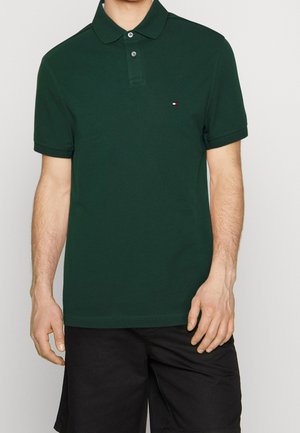 Mann trägt ein dunkelgrünes kurzärmeliges Poloshirt mit Knöpfen und schwarze Shorts, steht vor einem einfarbigen hellen Hintergrund.