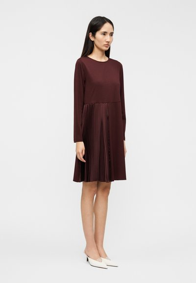 Μπορντό φόρεμα με μακριά μανίκια, που διαθέτει πλισέ φούστα. Ομαλή υφή στο πάνω μέρος, σε αντίθεση με τις δομημένες πτυχές από κάτω.