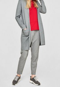 Cardigan lungo grigio chiaro sopra una maglietta rossa, abbinato a pantaloni grigi chiari con fantasia e sneakers scure. Sfondo neutro, attenzione ai dettagli dell'outfit.