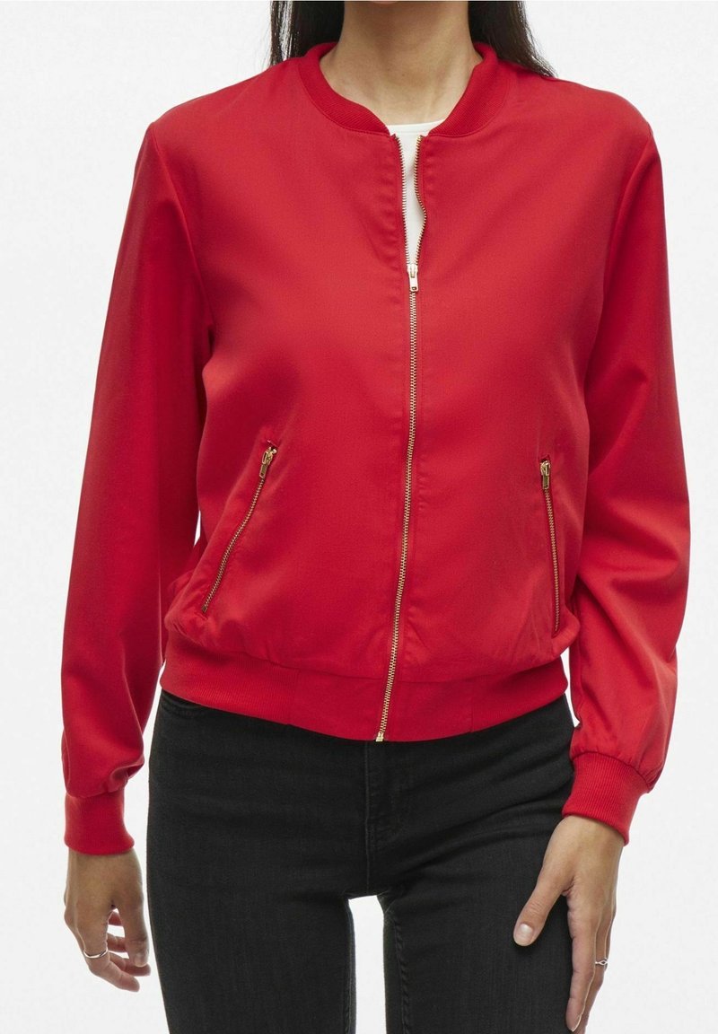 Femme portant une veste rouge vif à fermeture éclair, manches longues, fermetures éclair dorées et pantalon noir. Le haut du corps est visible, face vers l'avant.
