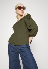 Blusa de ombros à mostra em verde-azeitona texturizada com detalhes de volantes e laços, combinada com calças de ganga de perna larga em azul-escuro. Óculos de sol oversized em castanho.