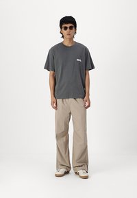 Camiseta de algodón gris con mangas cortas y un pequeño logo, combinada con pantalones beiges de corte holgado y zapatillas blancas. Lleva gafas de sol redondas.