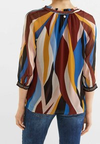 Blusa colorata con un motivo ondulato in blu, rosso, giallo e rosa. Tessuto semi-trasparente con scollatura a volant e polsini ricamati.