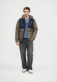 Jovem em pé, vestindo uma jaqueta bege e azul-marinho com padrão, uma t-shirt gráfica azul, calças cinzas largas e sneakers brancos, em fundo liso.