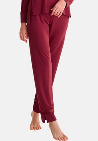 Pantalons lounge bordeaux avec une texture lisse, ornés de détails en dentelle à l'ourlet et d'une coupe décontractée. Pas de poches visibles. Pieds nus.