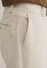 Pantalon beige sur mesure avec poche latérale fendue et passants de ceinture, porté avec un top en maille texturée marron rentré à l'intérieur.