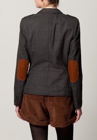 Femme portant un blazer écossais sombre avec des patches en cuir marron aux coudes et un short en velours côtelé marron, vue de dos contre un fond uni.