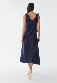 Maksimekolle navy sininen, jossa on röyhelökoristeet olkapäillä ja sivussa, valmistettu teksturoidusta kankaasta, ominaisuuksiin kuuluu V-mallinen selkäosa ja vartalonmyötäinen siluetti.