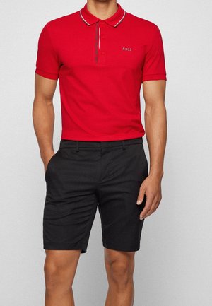 Mężczyzna w czerwonej koszulce polo BOSS z krótkim rękawem i czarnych szortach, stojący z rękami w kieszeniach na jasnoszarym tle.
