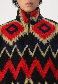 Fleece pullover with a textured surface featuring a geometric pattern in red, navy, black, and beige. High collar with a zip closure.