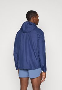 Navy blue lightweight jacket with a hood, featuring a smooth texture and a minimalist design. Includes a white accent on the arm.