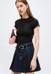 Νεαρή γυναίκα που φοράει εφαρμοστή μαύρη μπλούζα, πλισέ σκουρόχρωμη φούστα τζιν με μαύρη ζώνη, κρατώντας μια μικρή σκουρόχρωμη κόκκινη τσάντα σε λευκό φόντο.