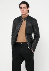 Μαύρο δερμάτινο μπουφάν με φερμουάρ μπροστά, ψηλό γιακά, εφαρμοστή γραμμή. Φορεμένο πάνω από μπεζ πουλόβερ, συνδυασμένο με μαύρο παντελόνι.