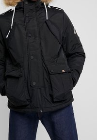 Svart puffjacka med foder i päls, flera framfickor, dragkedja och knappstängning samt vita dragskor. Texturerat tyg med en elegant design.