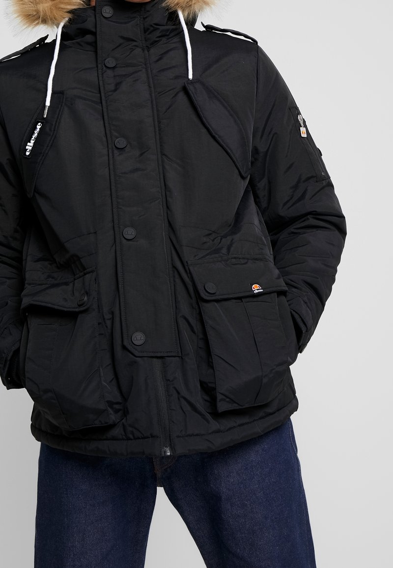 Svart puffjacka med foder i päls, flera framfickor, dragkedja och knappstängning samt vita dragskor. Texturerat tyg med en elegant design.