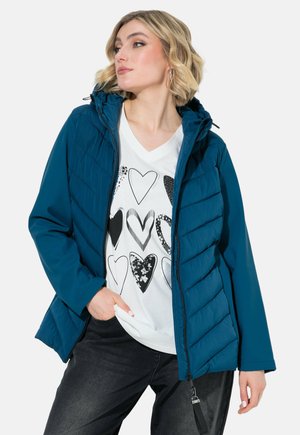 Ung kvinde iført en teal quiltet jakke over en hvid skjorte med sort-hvide hjerte-designs, poserer med den ene hånd i lommen.