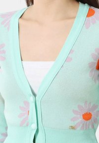 Cardigan verde menta con motivi floreali in rosa e arancione, scollatura a V, chiusura a bottoni e texture a coste. Accent rosa in oro sull'orlo.