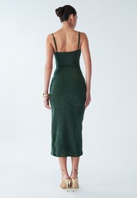 Eng anliegendes, dunkelgrünes Midikleid mit dünnen Trägern, glatter Textur und einem runden Rückenausschnitt. Gestylt mit zarten, hautfarbenen High Heels mit Riemchen.