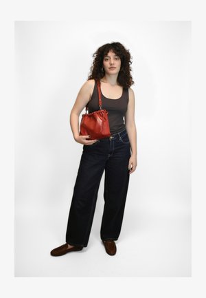 Geantă roșie din piele cu șnur de strângere și textură netedă; asortată cu blugi largi, cu talie înaltă, de culoare închisă și pantofi maro loafer. Design simplu și funcțional.