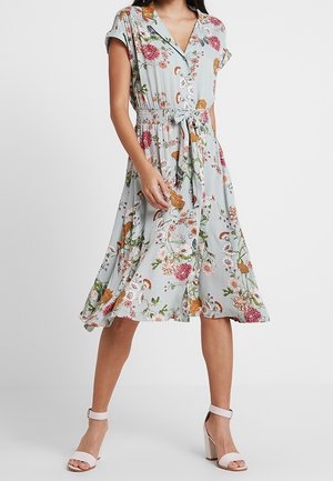 Frau trägt ein hellblaues Midikleid mit Blumenmuster, kurzen Ärmeln, Knopfleiste vorne, Taillengürtel und hellrosa Blockabsatz-Sandalen.