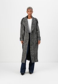 Lungo cappotto grigio a spina di pesce con collo oversize, tasche frontali e maniche arrotolate, abbinato a jeans a zampa scura e una maglietta bianca.