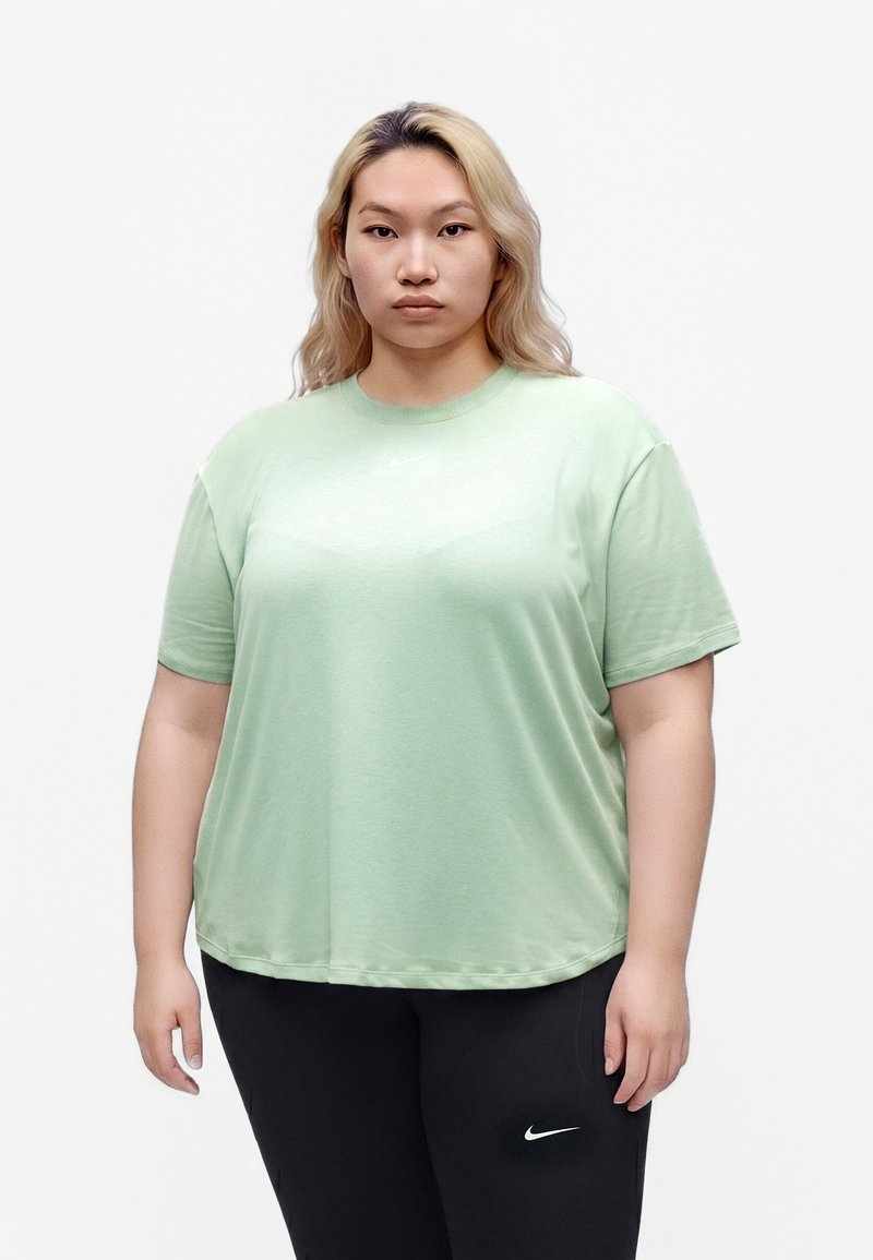 Femme aux cheveux blonds clairs portant un t-shirt vert clair Nike et un pantalon noir Nike, debout devant un fond blanc uni.