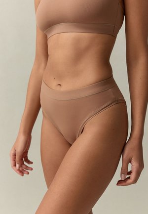 Vrouw draagt naadloze beha in huidskleur en bijpassend hoog uitgesneden ondergoed tegen een effen lichte achtergrond.