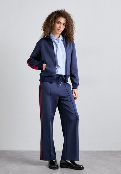 Giacca sportiva blu navy con strisce rosse, design con zip; abbinata a pantaloni jogging blu navy con strisce laterali rosse. Maglietta blu sotto.