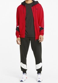 Rode zip-up hoodie met zwarte accenten, gecombineerd met zwarte joggers met witte diagonale strepen en witte sneakers met blauwe en rode details.