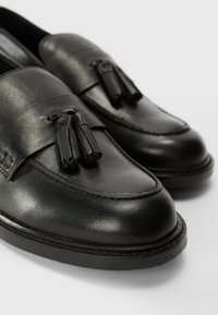 Mocassins en cuir noir avec des pampilles, bout rond, détails de couture et semelle en caoutchouc plate. Texture lisse et finition polie sont évidentes.