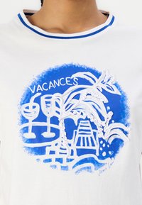 Hvid t-shirt med et blåt grafisk design med tropiske motiver, briller og en pyramide, omridset i hvidt. Blå ribbet halsudskæring.