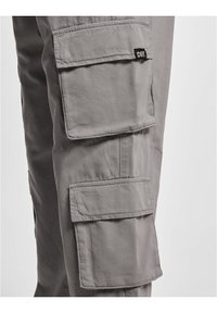Pantalon cargo gris avec plusieurs poches latérales à rabat, en tissu résistant, présentant un design avec coutures et une petite étiquette de marque.